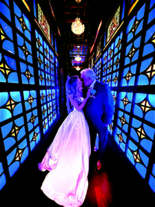 Bride and groom kiss beneath glowing chandeliers in a dramatic blue-lit corridor lined with stained-glass-inspired panels at their masquerade-themed wedding celebration.