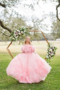 Woman in pink floral gown stands on grass before hexagonal flower arch, trees and field behind at Louisiana wedding.