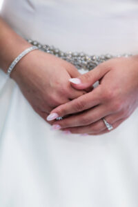 Hands with wedding ring and bracelet, fingers intertwined, in a white dress with jeweled waistband—wedding inspiration.