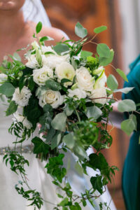 Bride holding a lush cascading white rose bouquet with greenery; dress and person in green visible, New Orleans wedding.