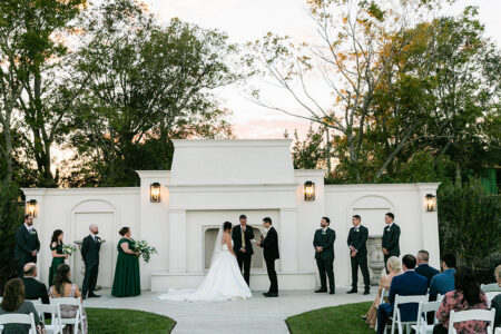 Bride and groom at outdoor altar with bridal party, guests on white chairs, trees, and sunset—Gulf South wedding inspiration.
