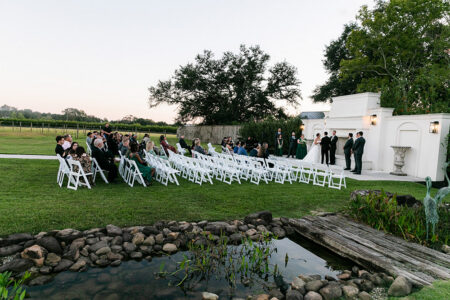Outdoor Gulf South wedding with guests on white chairs, green lawns, trees, pond, and a white ceremony structure.