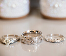 Close-up of engraved silver wedding bands and diamond engagement ring with intricate scrollwork details displayed on a reflective surface.