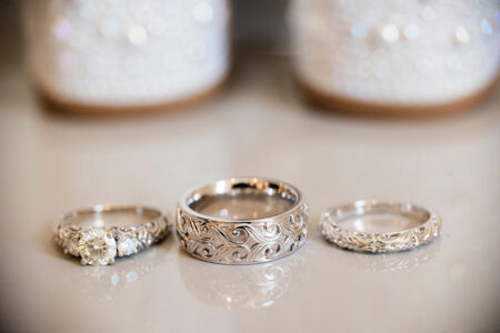Three elegant silver rings—engraved, diamond, and patterned—on a reflective surface with sparkling blurred background.