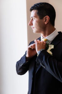 Man in black suit and yellow tie adjusts collar by a bright window, white flower on lapel—stylish Louisiana wedding inspiration.