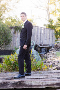 Man in black suit with white boutonniere stands on garden bridge, surrounded by greenery, fence, and bird statues at Louisiana wedding.