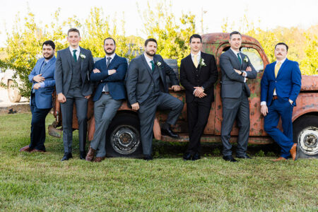 Seven groomsmen in suits pose by a rusty truck on grass at a relaxed Louisiana wedding, with trees and clear skies behind them.