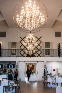 Bride in white gown enters elegant hall with chandeliers, walking down aisle with suited man as guests watch.