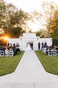 Louisiana sunset wedding outdoors with guests, couple at altar, attendants, white backdrop, and green lawn.