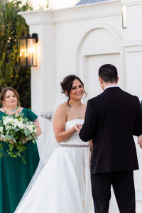 Bride in white gown smiles at groom during outdoor Louisiana wedding as bridesmaid in green dress with bouquet stands nearby.