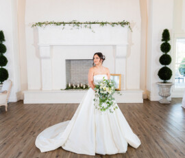 Bride in a strapless satin ball gown posing with a cascading white and greenery bouquet inside a bright white reception hall with a grand fireplace backdrop.