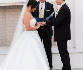 Bride and groom exchanging rings during an outdoor wedding ceremony as the officiant reads from a blue binder beside them.