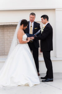 Bride and groom hold hands at Gulf South wedding as officiant reads; bride in white gown and veil, groom in black suit.