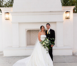 Newlyweds posing in front of a white outdoor fireplace feature, the bride holding a lush greenery bouquet with her satin train flowing across the ground.