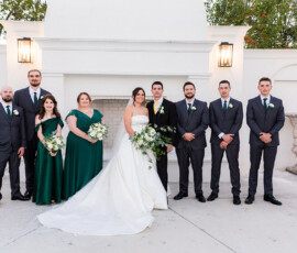Wedding party portrait with the bride and groom surrounded by bridesmaids in emerald green dresses and groomsmen in dark suits outside the ceremony space.