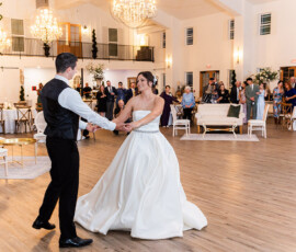 Bride and groom spinning across the dance floor during their first dance beneath glowing chandeliers in an elegant white reception hall.