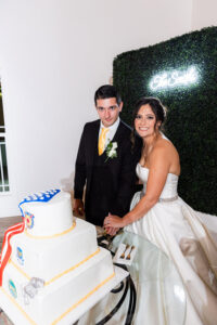 Bride and groom smile cutting a tiered cake at their Louisiana wedding, with neon sign and greenery backdrop.