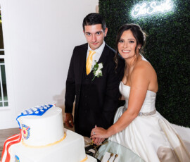 Newlyweds cutting a white tiered wedding cake decorated with military-inspired emblems beside a greenery wall and neon sign at the reception.