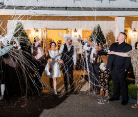 Bride and groom making a festive streamer exit at the end of the reception while guests celebrate around them outdoors at night.