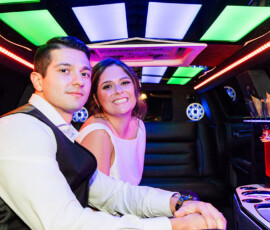 Newlyweds sitting together inside a limousine with colorful neon lighting and champagne glasses after their wedding reception.