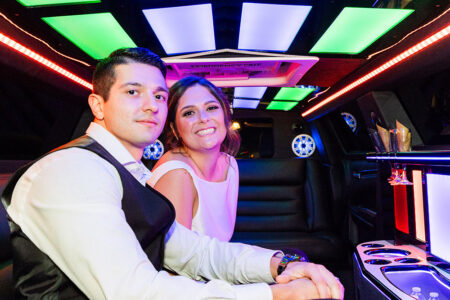 Bride in white dress and man in vest smile together in neon-lit limo, celebrating their New Orleans wedding with drinks.