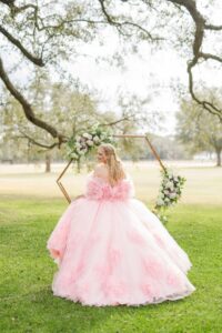 Woman in voluminous pink gown stands on grass, looking back by floral arch and trees at a Louisiana wedding under bright sky.