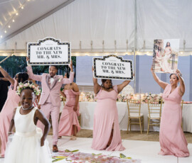 Wedding party in pink holds “Congrats to the New Mr. & Mrs.” signs under a white tent as a smiling flower girl walks by.