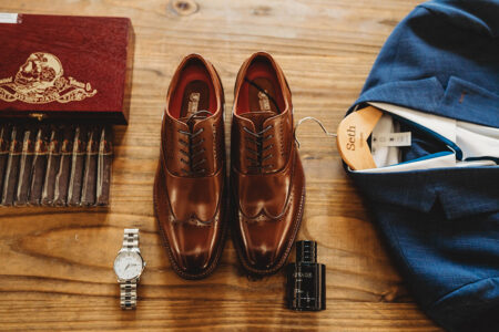 Brown dress shoes, blue suit, silver watch, black cologne, and cigars arranged on wood for Gulf South wedding inspiration.