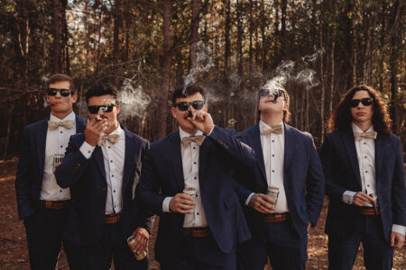 Five men in blue suits and bow ties enjoy drinks and cigars outdoors, embodying effortless wedding style and camaraderie.