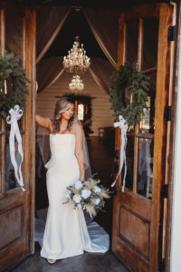 Bride in white dress and veil stands in flower-adorned doorway with chandeliers inside, perfect New Orleans wedding inspiration.