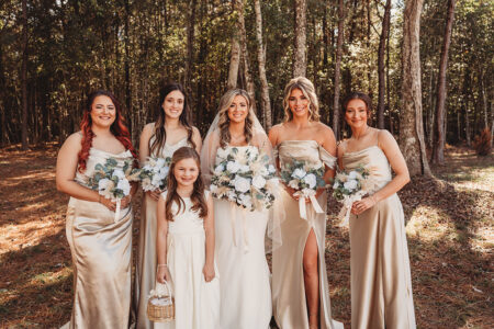 Bride and bridesmaids in gold dresses smile outdoors, with a flower girl holding a basket at a sunny Louisiana wedding.