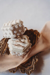 Gold rings with a diamond in a pearl-accented hexagon box on a gold tray and pink fabric at a Louisiana wedding.
