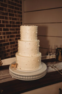 Elegant three-tiered white wedding cake with pearls and a dog figurine, flanked by champagne flutes and forks.