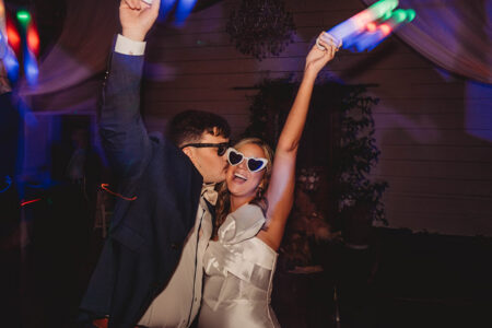Couple joyfully dances at a Louisiana wedding, woman in heart sunglasses and white dress, smiling under colorful party lights.