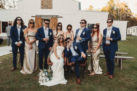 Outdoor wedding party with bride and groom in sunglasses, bridesmaids in gold, groomsmen in blue, all posing with drinks.