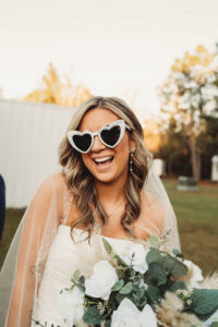 Bride in pearl heart sunglasses holds white bouquet outdoors with trees and a white building in the background.