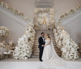 Bride and groom hold hands on a grand, flower-lined staircase at an elegant New Orleans wedding reception.