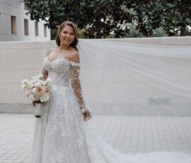Bride in long-sleeved lace gown with veil, holding white roses and smiling outdoors at her Gulf South wedding.