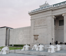 White-clothed cocktail tables stand outside a grand stone building under a pink sky, perfect for a dreamy New Orleans wedding.