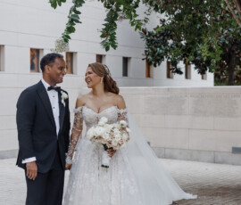 Bride in lace dress and groom in black tux smile outdoors under a leafy tree at their classic New Orleans wedding.