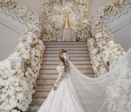 Bride in white gown on flower-adorned stairs faces gold wedding cake under MR & MRS sign at a classic New Orleans wedding.