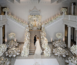 Bride and groom on a flower-lined staircase in an elegant hall with white floral decor, evoking a classic New Orleans wedding.