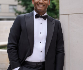 Smiling man in black tuxedo leans against a wall outdoors before a Louisiana wedding, with trees and building behind him.