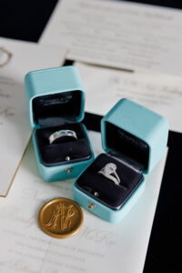 Tiffany & Co. wedding bands and pear-shaped diamond engagement ring displayed in blue velvet boxes atop formal wedding invitation suite