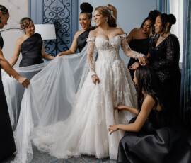 Bride in white gown smiles as six women in black dresses adjust her dress and veil in a softly lit, blue-accented room.
