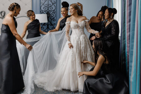 Bride in long-sleeve floral lace wedding gown surrounded by bridesmaids in black satin dresses while preparing for ceremony