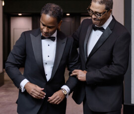Two men in black tuxedos share a supportive moment indoors before a Louisiana wedding, surrounded by elegant decor.