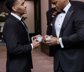 Two men in tuxedos face each other indoors holding blue boxes, as someone adjusts a collar at a joyful Louisiana wedding.