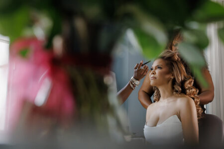 Bride having makeup applied near hotel window with soft curls and strapless getting-ready outfit reflected through greenery foreground