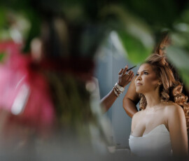 Bride in white dress has her hair styled, gazing thoughtfully out window with blurred foliage in foreground.